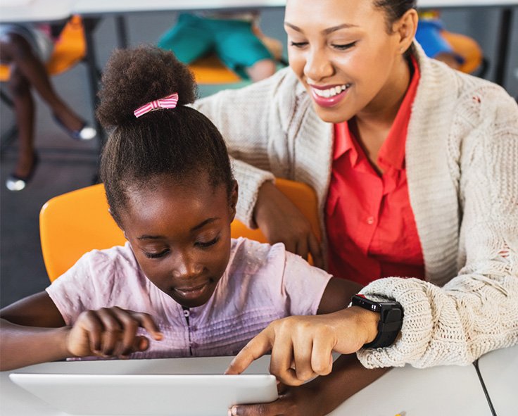 A teacher in a classroom, guiding a young child through a classroom resource on a tablet device