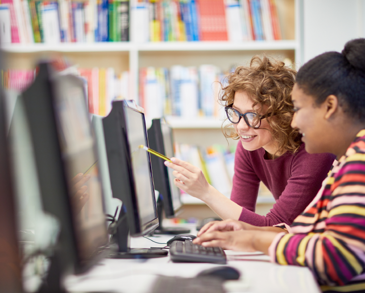 A teacher pointing out a detail in a student's coursework to the student, using Snapplify on a desktop computer in a library